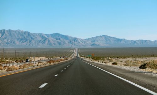 Empty road amidst field leading towards mountains against clear blue sky