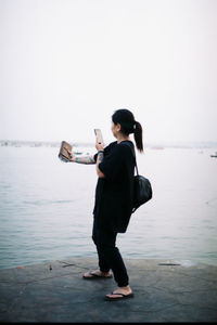 Woman photographing book with mobile phone on shore against clear sky