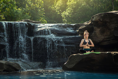 Teenage girl with hands clasped meditating while sitting on rock in lake at forest