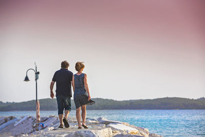 Rear view of couple walking on pier by sea against clear sky