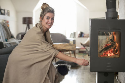 Portrait of smiling woman putting wood in fireplace at home