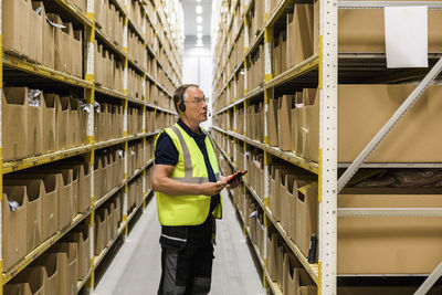 Side view of senior male worker with digital tablet looking at packages on rack while talking through headset at distrib