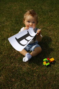 High angle view of baby boy holding paper with letter e while sitting on grassy field at park