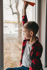 A boy with an orange paper airplane, sits at the window and looks out into the street.