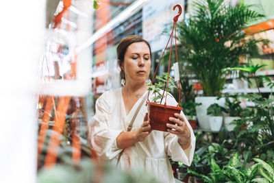 Brunette middle aged woman in white dress buys green potted house plants at garden store