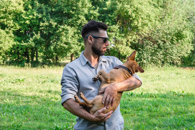 Handsome stylish european man is holding his dog in hands in park on a walk.