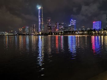 Illuminated buildings by river against sky at night