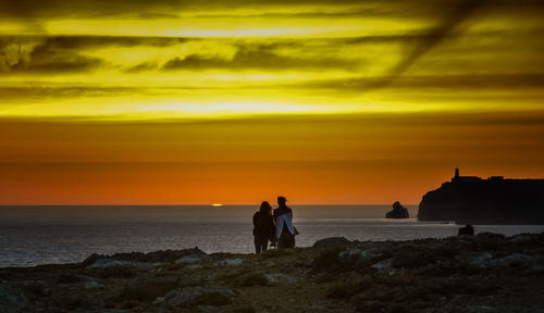 Rear view of friends standing at sea shore against orange sky