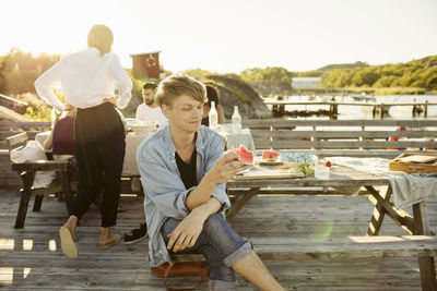 Smiling man eating watermelon slice while friends sitting in background at jetty