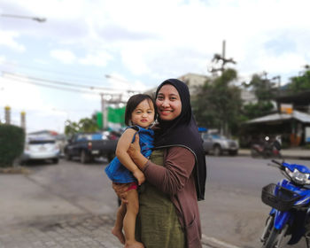 Portrait of smiling mother carrying daughter while standing on street in city