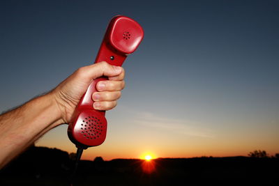 Close-up of hand holding red telephone receiver against sky during sunset