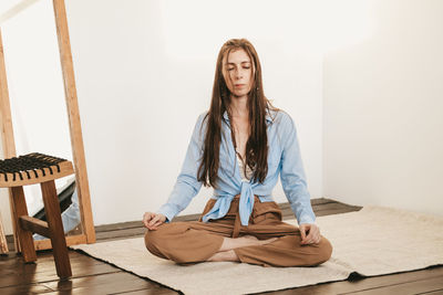 Girl with long and dark hair sitting lotus pose meditating on floor light room interior. 