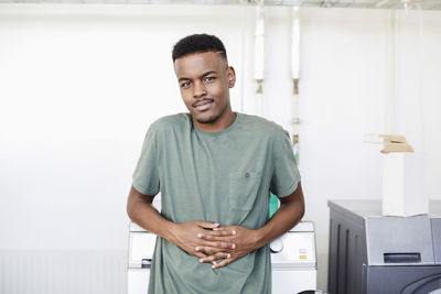 Portrait of young man with hands clasped standing by washing machine