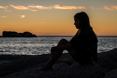 Silhouette woman sitting at beach against sky during sunset