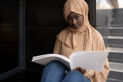 Crop concentrated young african american muslim female student in traditional headscarf sitting on street and reading book while preparing for exams