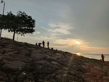 People on rocks at shore against sky during sunset