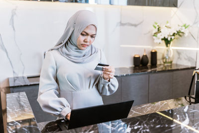 Mid adult woman wearing hijab doing online shopping over laptop while standing at home