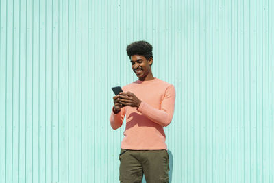 Young man using smart phone while standing against wall