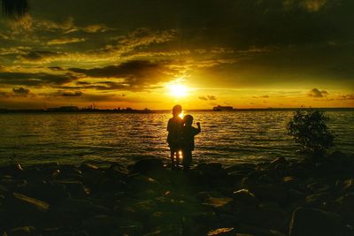 Silhouette brothers standing at beach against sky during sunset