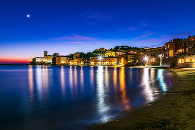 Illuminated buildings by river against sky at night