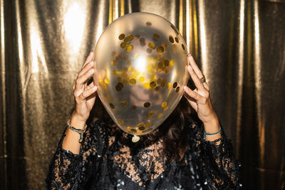 Unrecognizable female wearing festive dress covering face with air balloon with golden confetti while standing on golden background at party