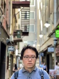 Portrait of young asian man in eyeglasses standing in laneway against buildings.