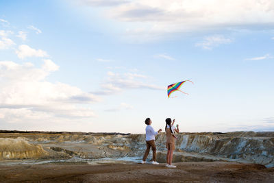 A boy and a girl launch a bright kite into the sky in the mountains near the river.