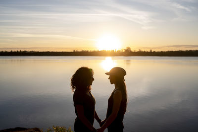Lesbian couple looking at each other and hugging while standing by a lake at sunset.