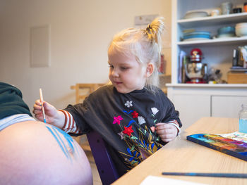 Cute girl painting on pregnant mother's abdomen at table in kitchen