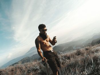 Shirtless mid adult man using mobile phone while standing on mountain against sky