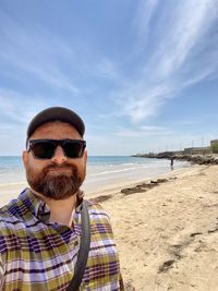 Portrait of young man wearing sunglasses while standing at beach against sky