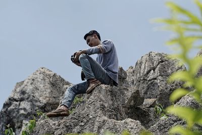 Low angle view of young man sitting on rock against clear sky
