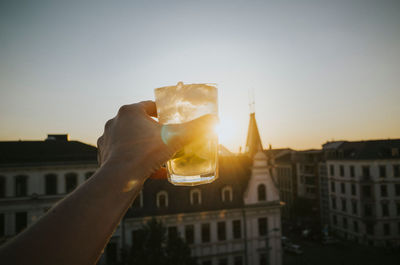 Cropped image of hand holding drink against buildings during sunset