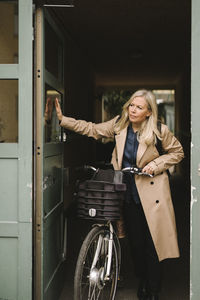 Businesswoman wearing long coat opening door while holding bicycle