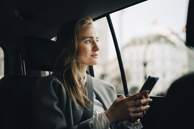 Contemplating female entrepreneur with smart phone looking through window while sitting in taxi