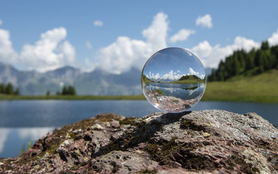 Reflection of crystal ball on rock by lake against sky