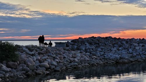 People on rocks by sea against sky during sunset