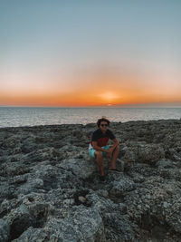 Man sitting on rock at beach against sky during sunset