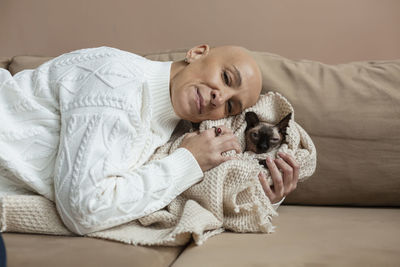 Bald woman embracing sphynx cat wrapped in blanket on sofa