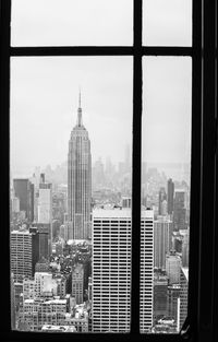 Empire state building in city against sky seen through glass window