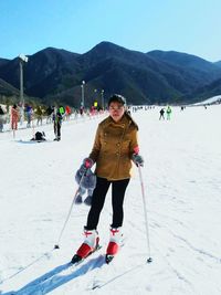 Portrait of woman wearing ice skates holding ski poles on snow covered mountain against clear sky