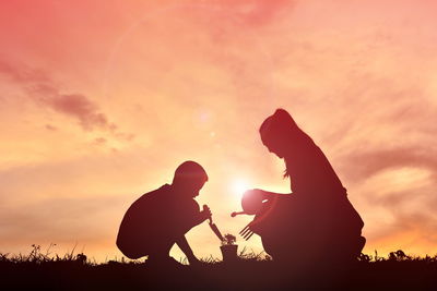 Silhouette siblings planting on field against sky during sunset