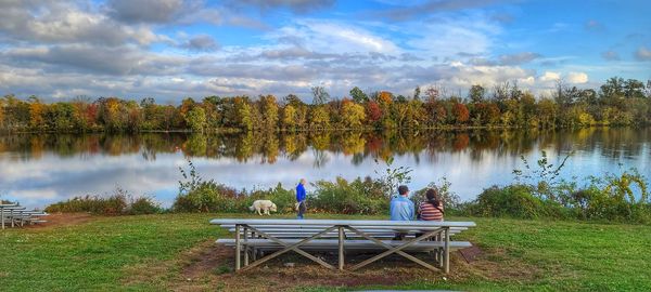 Panoramic view of man sitting on grass by lake against sky