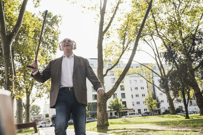 Happy senior man with walking cane listening to music through wireless headphones and dancing on footpath