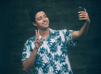 Smiling young man gesturing while taking selfie through mobile phone