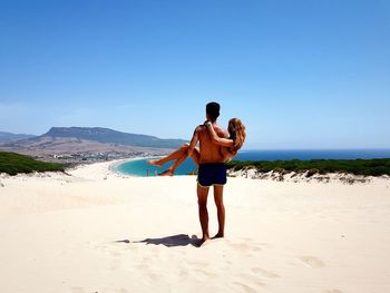 Rear view of shirtless man lifting girlfriend while standing at beach against clear sky