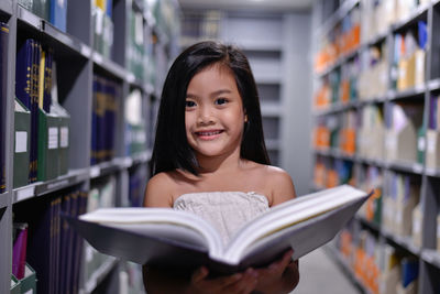 Portrait of cute smiling girl holding book while standing amidst bookshelves in library