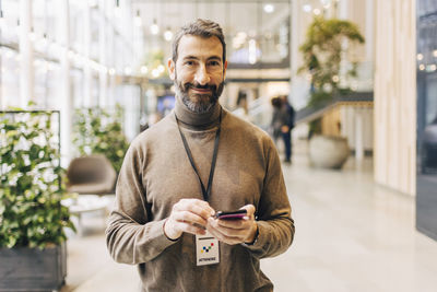 Portrait of smiling mature businessman holding smart phone while standing at convention center
