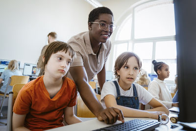 Smiling female teacher assisting girl with down syndrome in computer class at school