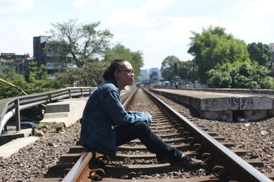 Side view of young man sitting on railroad track against sky